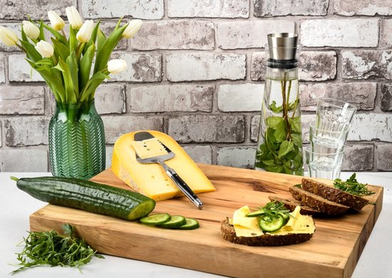Gouden kaas op een houten plank met komkommer, boterhammen en witte tulpen in een groene vaas.
