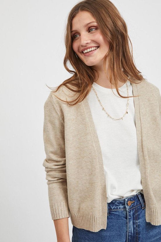Een lachende vrouw draagt een beige vest over een wit shirt en jeans.