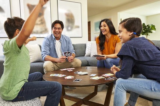Familie speelt kaartspel aan ronde tafel, man in blauw lacht en houdt kaarten vast, vrouw in oranje, jongen in groen, meisje in blauw.