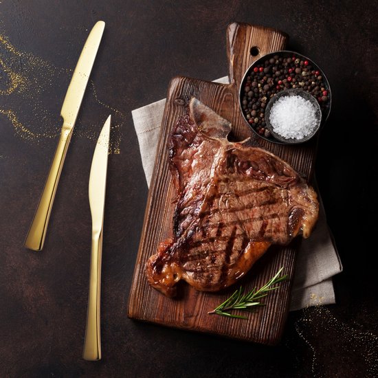 Gegrilde T-bone steak op een houten plank met rozemarijn, peperkorrels en grof zout, naast twee gouden messen.