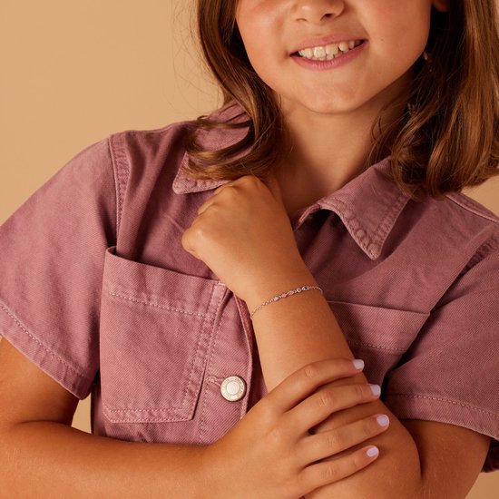 Een jong meisje met een roze jurk draagt een zilverkleurig armbandje met gekleurde hartjes.