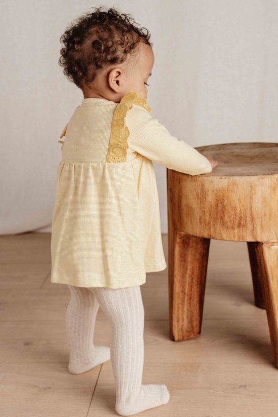 Baby in een gele geruite jurk met kanten schouderdetails, van achteren gezien.