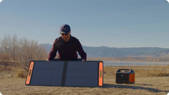 Een man met een pet houdt een Jackery zonnepaneel omhoog in een open veld, naast een oranje power station.