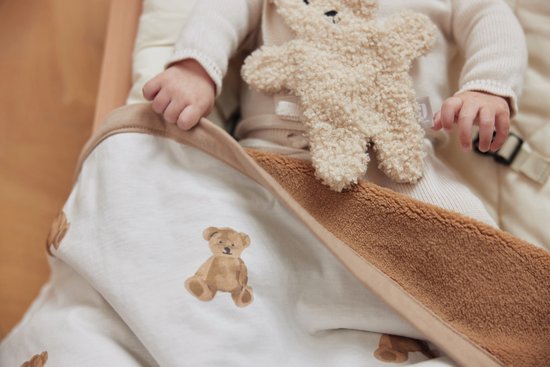 Bébé avec peluche ours sur une couverture blanche à imprimé ours en peluche et bordure marron, dans un berceau.