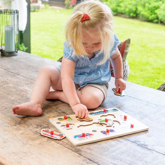 Een jong meisje met blonde paardenstaart speelt met een houten puzzel met voertuigen op een tafel.