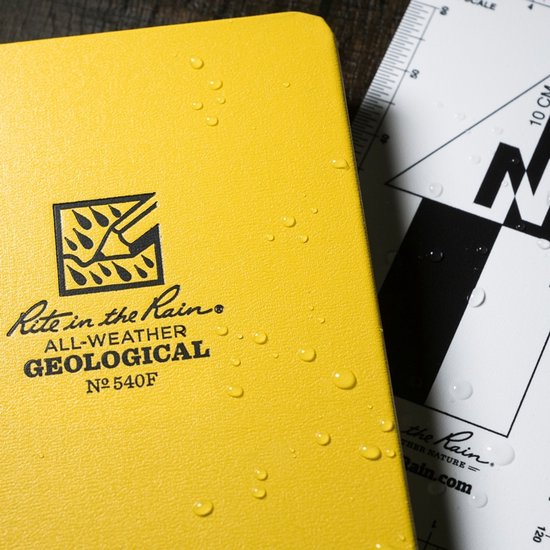 Gele kaft van 'Rite in the Rain ALL-WEATHER GEOLOGICAL' notitieboek, met waterdruppels. Rechts een deel van een meetlat.