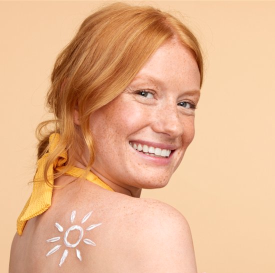 Vrolijk lachende vrouw met sproeten, zonnegevoelige huid, draagt gele bikini. Zonnecrème in zonvorm op haar rug.