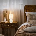 Witte tafellamp met geribbelde voet en witte stoffen kap op een houten nachtkastje.