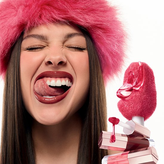 Model met roze bonten hoed likt met tong over glanzende, roze lippen. Stapel Lancôme lipgloss flesjes ernaast.