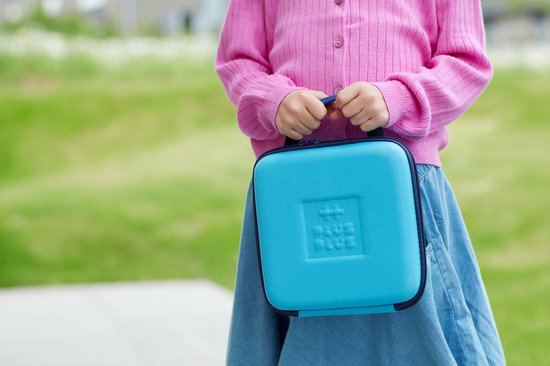 Een kind draagt een blauwe Plus-Plus reiskoffer met handvat in een groene, buitenomgeving.
