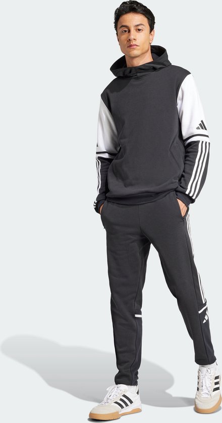 Man met een donkergrijs sweatshirt met witte mouwen en Adidas-strepen op de mouwen, staand.