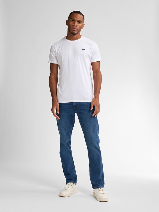 Man staat frontaal in een witte t-shirt met logo en een blauwe spijkerbroek, rechte pasvorm, met witte sneakers.