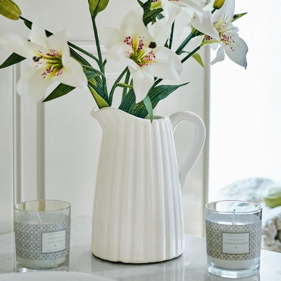 Witte karaf met verticale ribbels, met witte lelies in een vaas en twee geurkaarsen.