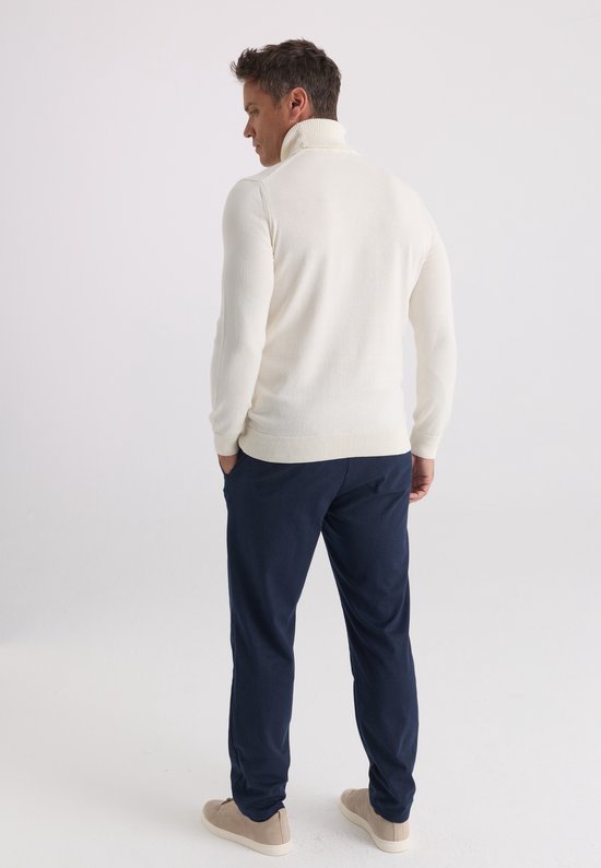 Aanzicht op de rug van een model in een ecru coltrui, donkerblauwe broek en beige sneakers tegen een witte achtergrond.