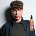 Jonge man met bruine krullen kijkt opzij naast een fles Reuzel Spray Grooming Tonic.