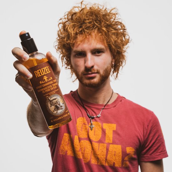 Man met rood krullend haar houdt een fles Reuzel Spray Grooming Tonic omhoog.