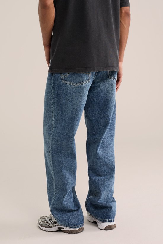 Achterkant van een man in een loszittende, blauwe spijkerbroek en grijze sneakers. De broek heeft twee achterzakken.