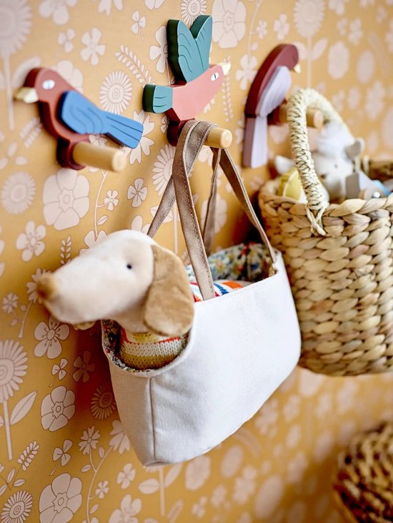 Een stoffen mandje met een witte hond en een rieten mand met speelgoed hangen aan een muur met bloemenbehang, aan houten vogelkapstokken.
