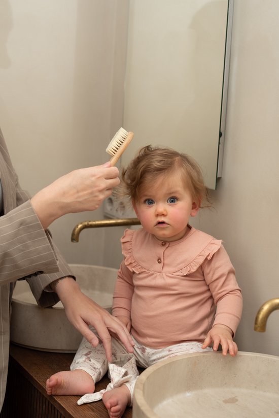 Baby in roze Dirkje kledingset zit op een wastafel terwijl haar haar wordt geborsteld.