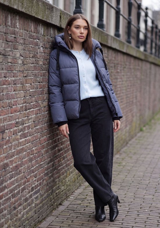 Model in een blauwe pufferjas, lichtblauwe cardigan en zwarte spijkerbroek, staand bij een bakstenen muur.