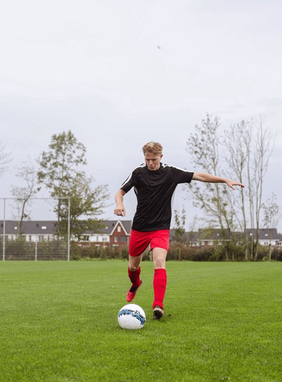 Jonge man in zwart voetbalshirt en rood voetbalbroekje trapt tegen een voetbal.