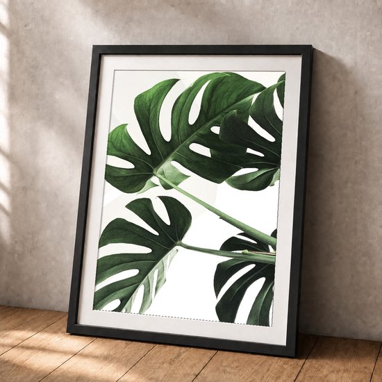 Een poster met groene monstera bladeren in een zwart frame, staand op een houten vloer met een betonnen muur.