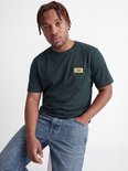 Man met gevlochten haar draagt een donkergroen T-shirt met geel logo op de borst.