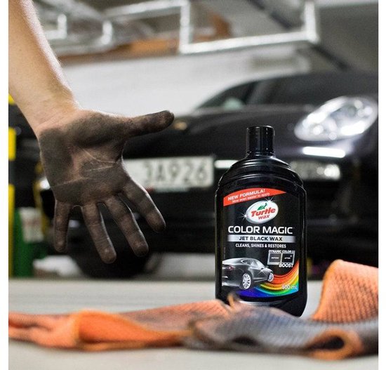 Hand met zwarte smurrie boven zwarte fles Turtle Wax Color Magic Jet Black Wax naast oranje doeken. Zwarte auto op achtergrond.