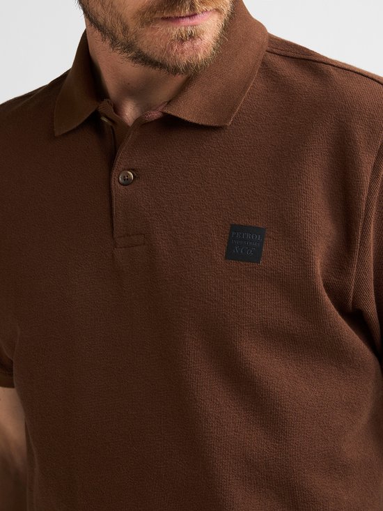 Close-up van een bruin katoenen poloshirt met een geborduurd logo op de borst en een knoopsluiting.