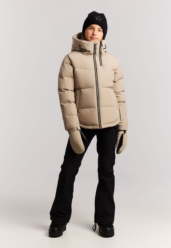 Een jong meisje met blonde haren, draagt een beige winterjas, zwarte muts, ski-handschoenen en skibroek.