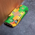 Een kokervormige kakkerlakkenval met groene, gele en oranje kleuren, met afbeeldingen van huizen en kakkerlakken. Geplaatst op een grijze vloer naast een houten deurpost.