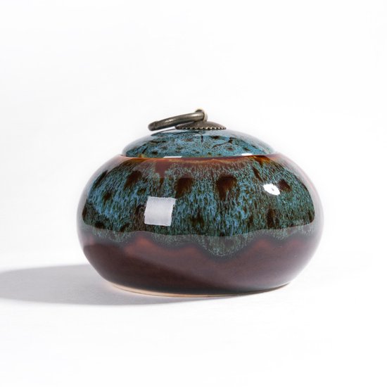 Ronde, keramische urn met deksel in blauwgroen en bruin met een metalen handvat.