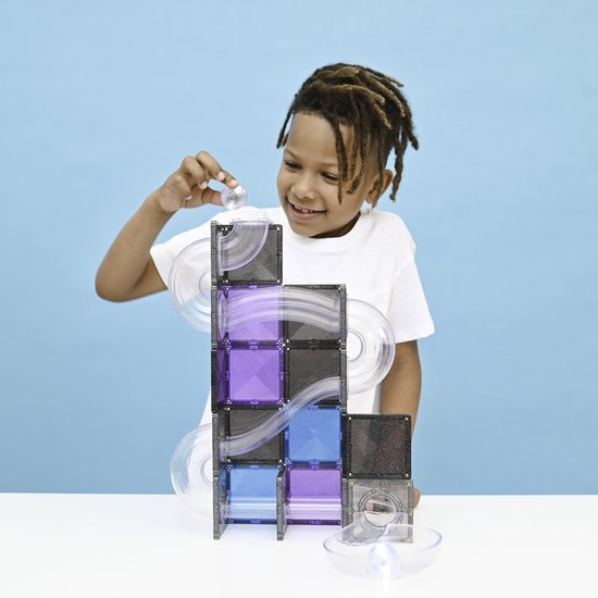 Een jongen met dreadlocks en witte T-shirt plaatst een knikker in een paarse Connetix knikkerbaan met blauwe en grijze tegels.