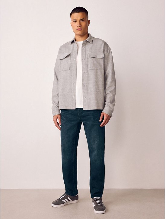 Model draagt een donkerblauwe stretch jeans, wit t-shirt, grijs overhemd en grijze sneakers.