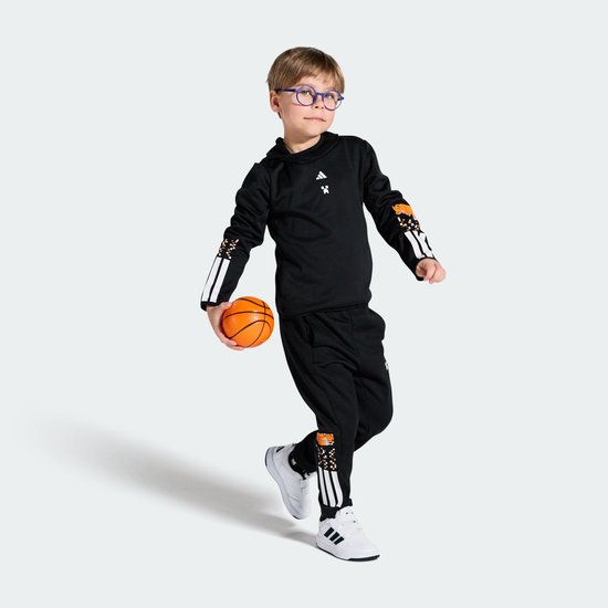 Jongen in zwart Adidas trainingspak met oranje grafische details op de mouwen en broekspijpen, en witte strepen. Hij houdt een basketbal vast.