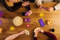 Een groep vrienden speelt Music Match aan een tafel met popcorn, drankjes en chips. Iedereen heeft kaarten in zijn handen.
