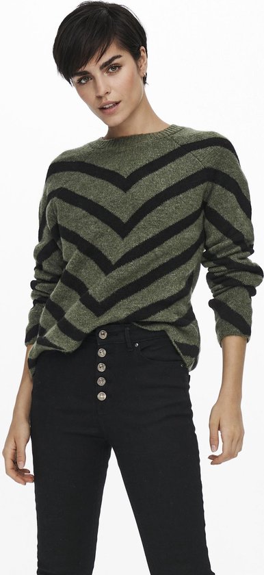 Model in olijfgroene trui met zwarte V-strepen en zwarte broek met knopen.