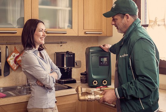 Man in groen uniform met monteur, naast een groene drukverhoger, praat met een vrouw in een keuken.