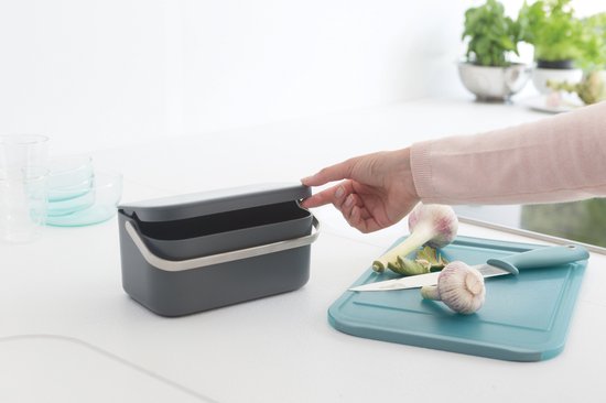 Vrouw opent donkergrijs afvalbakje in keuken met groenten ernaast.