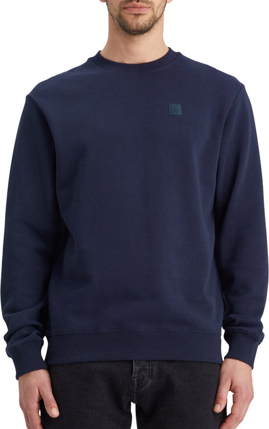 Man met donkerblauwe sweater met ronde hals en vierkant logo op de borst.