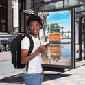Jonge man met koptelefoon en smartphone voor een bushokje met poster van oranje camper met surfboards.