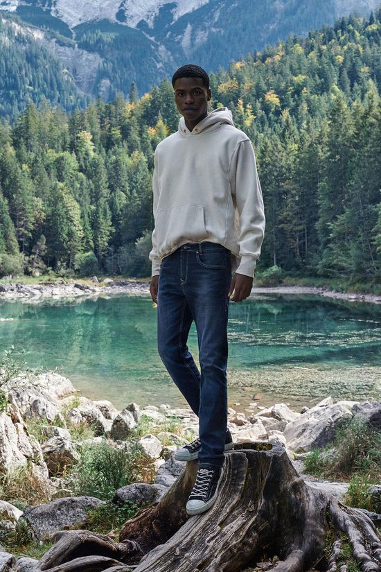 Man in beige hoodie en donkerblauwe jeans, staand op een boomstronk aan een meer.