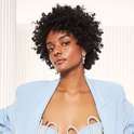 Model met natuurlijk krullend haar, draagt een lichtblauwe blazer en een top met parels.