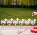 Zeven gevulde glazen met roze drank en limoenschijfjes, op een houten tafel buiten.