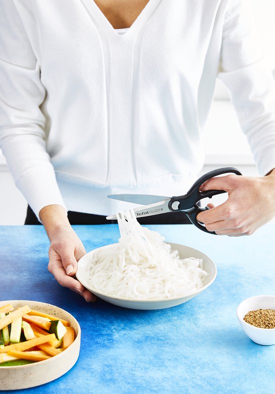 Zwarte keukenschaar snijdt witte rijstnoedels fijn in een witte kom.