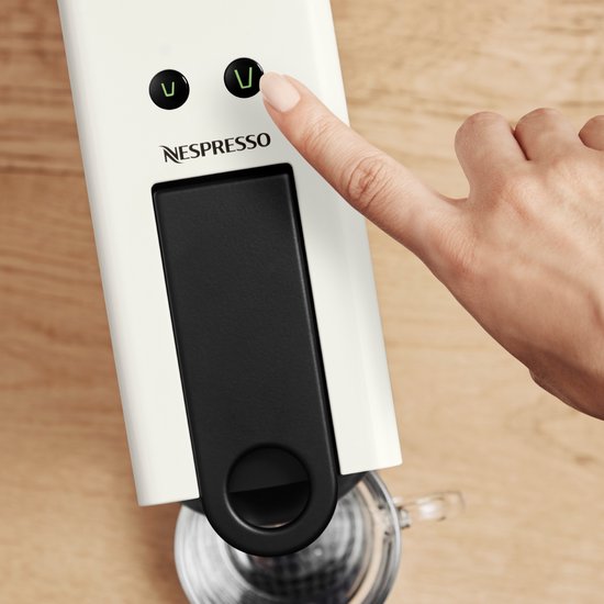 Un doigt appuie sur la machine Nespresso Essenza Mini blanche, avec des voyants verts.
