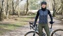 Vrouw met fiets in het bos, draagt een zwarte helm, nekomslagdoek, zwart/wit gemêleerd fietsshirt met lange mouwen en olijfgroene fietsbroek.