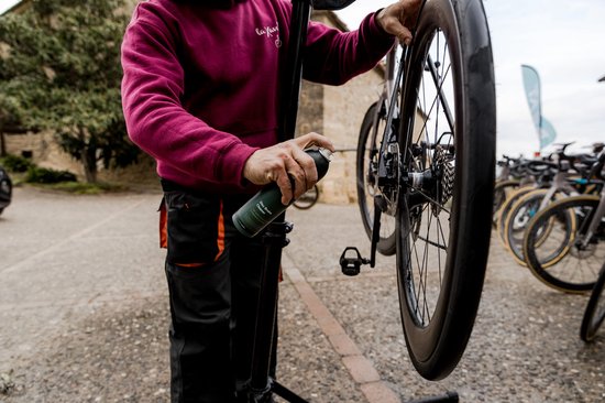 Persoon in bordeaux trui spuit remreiniger op een racefietswiel op een fietsstandaard.