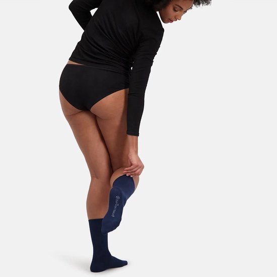 Een persoon trekt een donkerblauwe bamboe sok aan. De persoon draagt een zwarte slip en een zwart shirt met lange mouwen. De sok reikt tot net onder de knie.