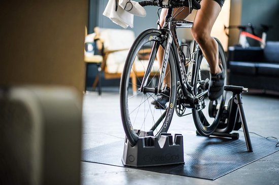 Een racefiets op een zwarte fietstrainer met het logo 'CycleOps', op een zwarte mat.